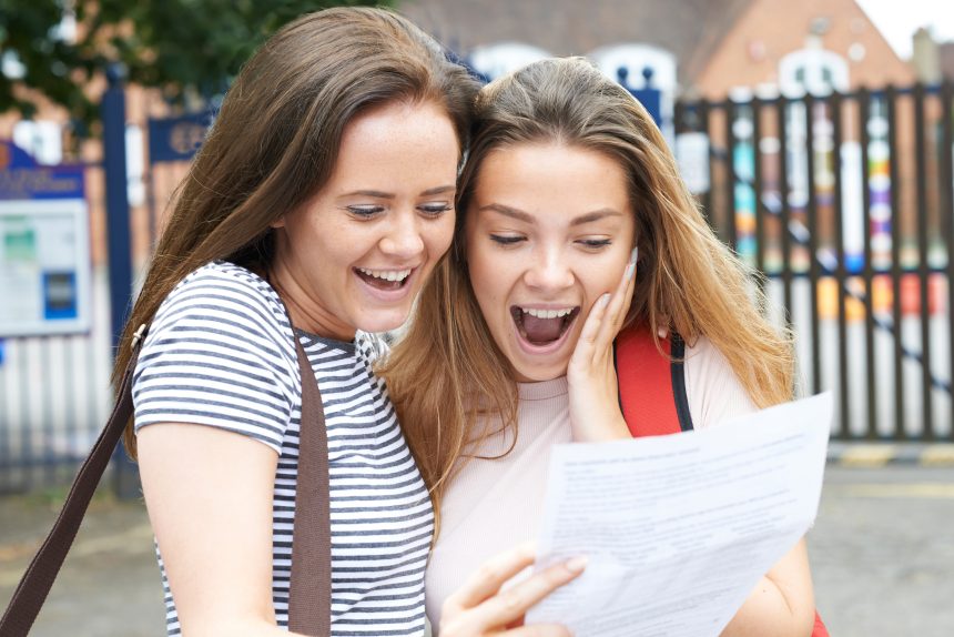 A happy and surprised teenage girl looking at her exam results on Results Day with her friend.