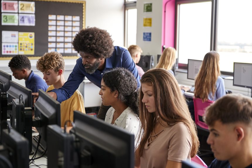 A male teacher helping sixth form students in A Level IT class.