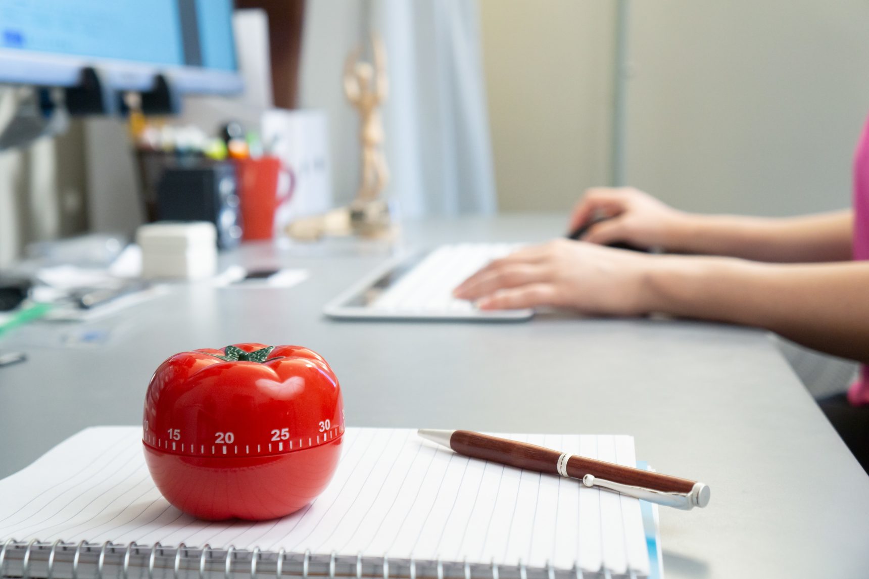 A Pomodoro tomato timer sits on a desk on top of a notebook with a student typing on a computer keyboard in the background.