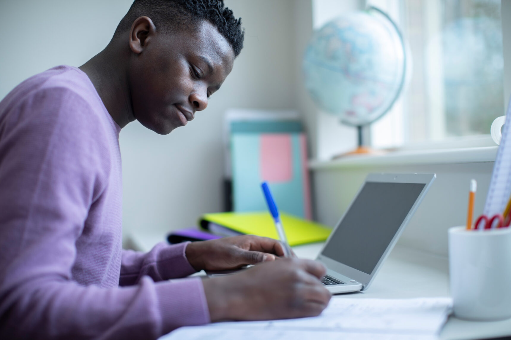 A teenage boy working at a desk in a home study space filled with natural light.