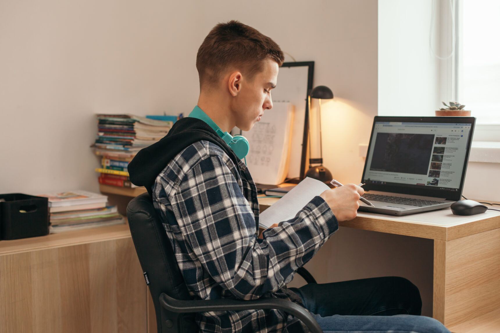 A teenage boy sitting at a desk and reading a book in a home study space with a desk lamp and a laptop.