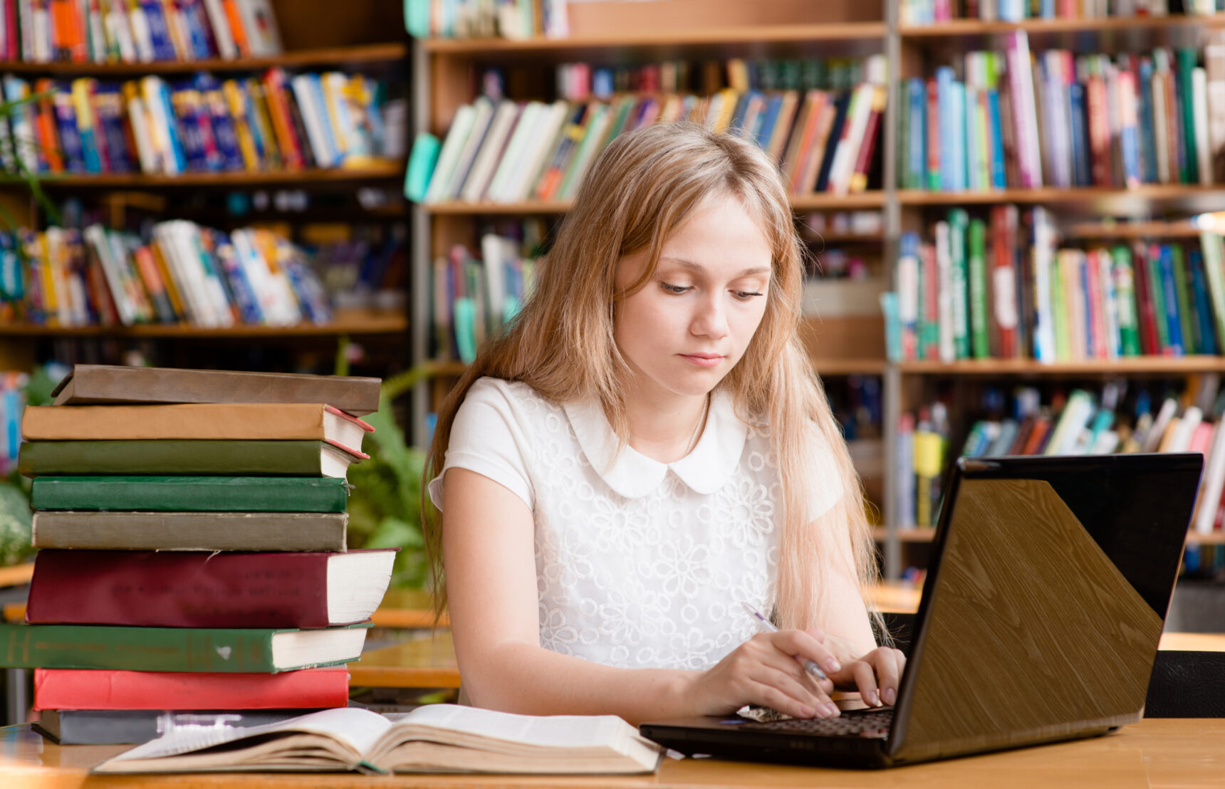 A teenage girl is studying in the library with a laptop surrounded by a pile of text books.