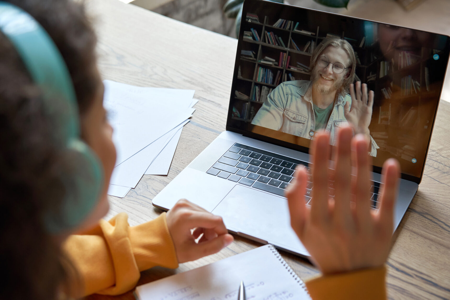 A female student wearing headphones sits in front of her laptop and waves to her online tutor.