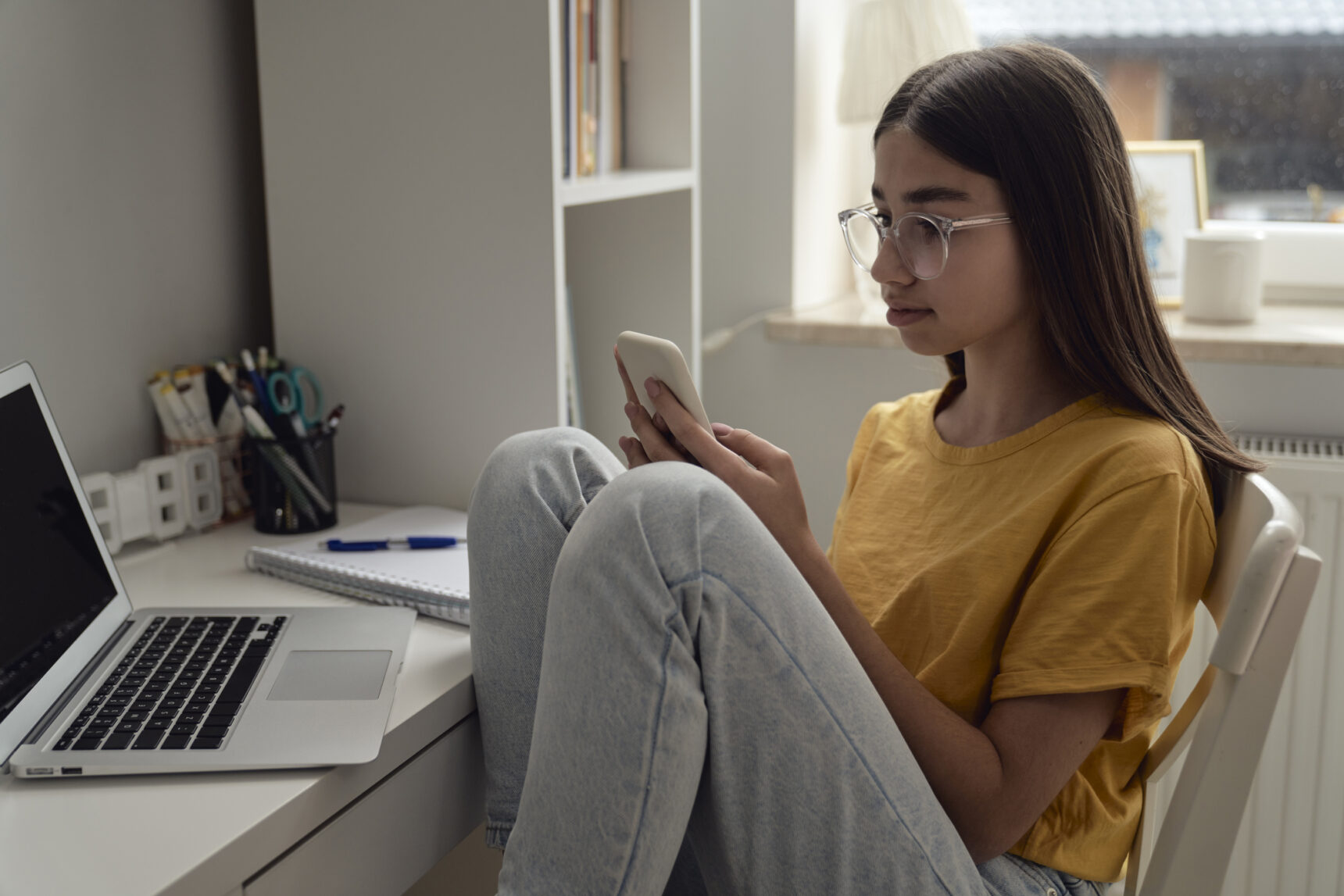A teenage girl is looking at her smartphone during a study session as she sits at her desk surrounded by a laptop and a notebook and pen.
