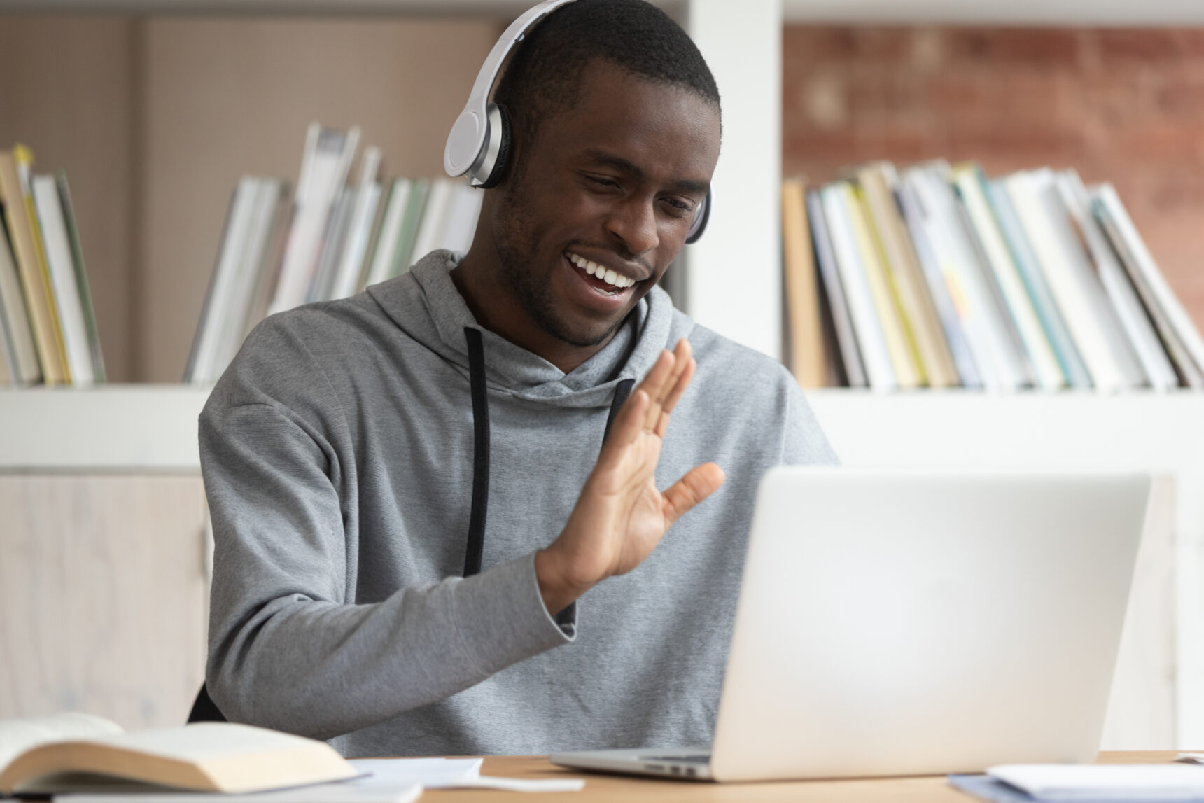 A male online tutor is smiling and waving at a student as he teaches a lesson via laptop.