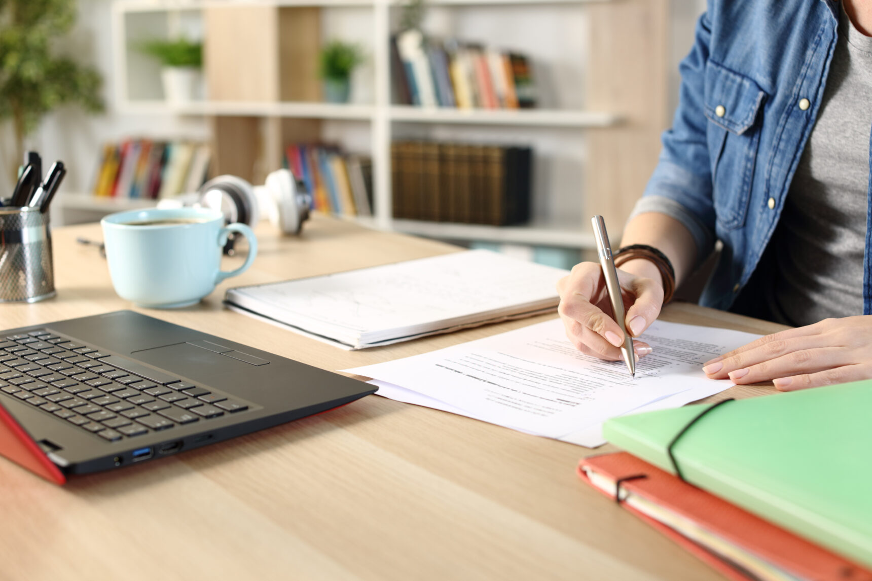 A tutor sits at a desk writing a list surrounded by folders and a laptop as they carry out admin tasks for their tutoring business.