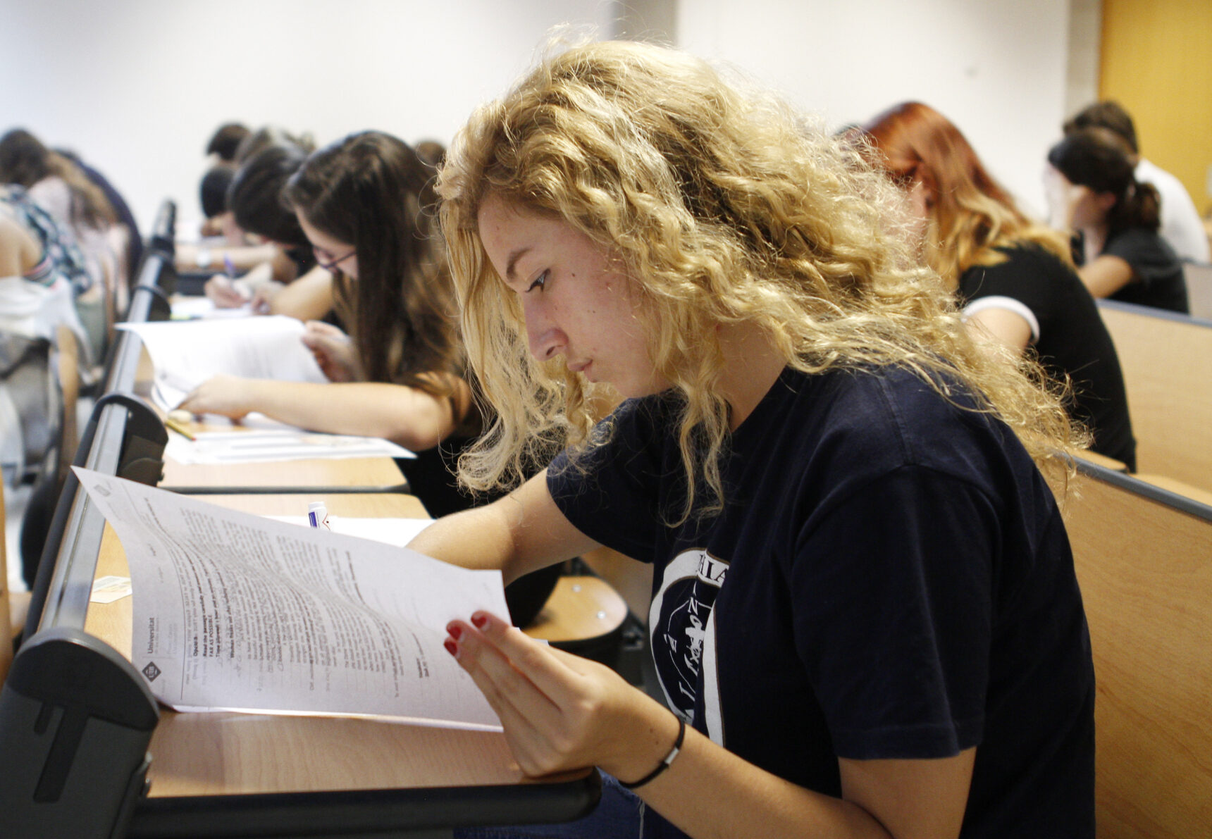 A female A Level student reading an exam paper as she sits at a desk in an exam hall.