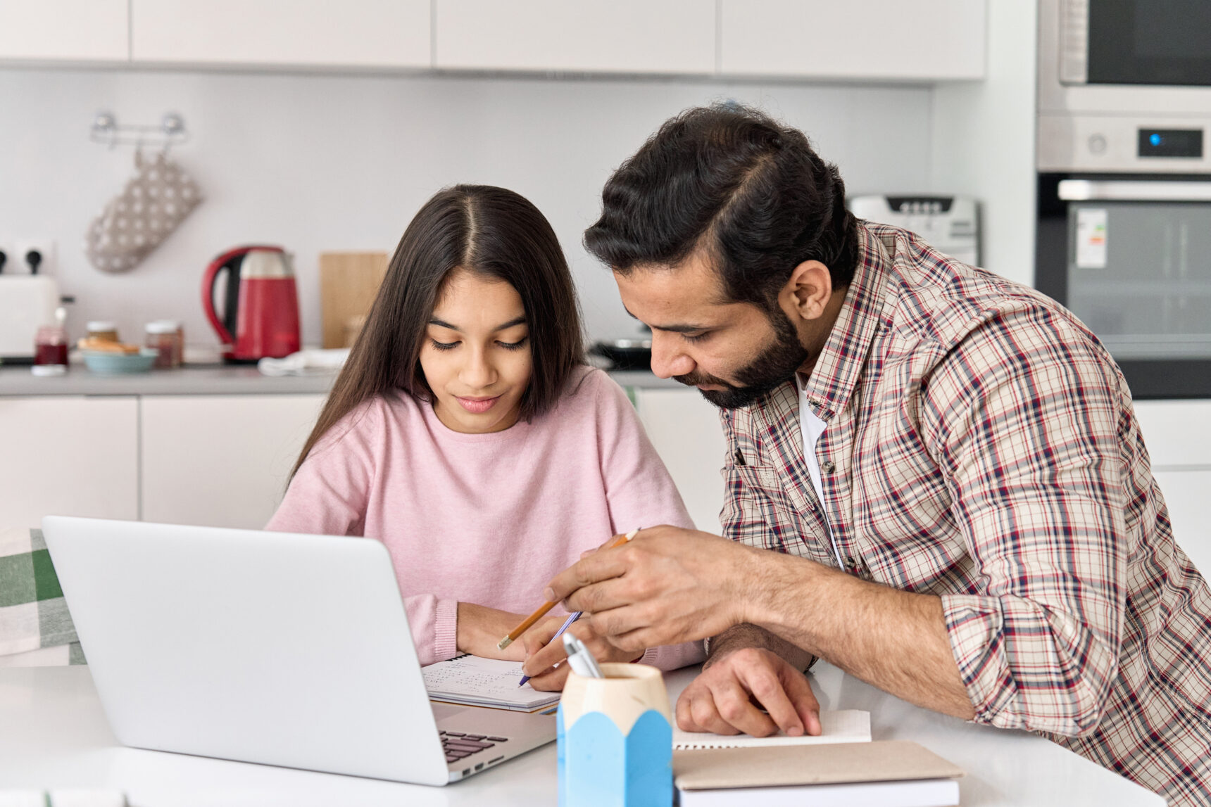 A Dad is sitting at the kitchen table with his daugther, helping her to prepare for her mock exams as she reviews notes in front of a laptop.