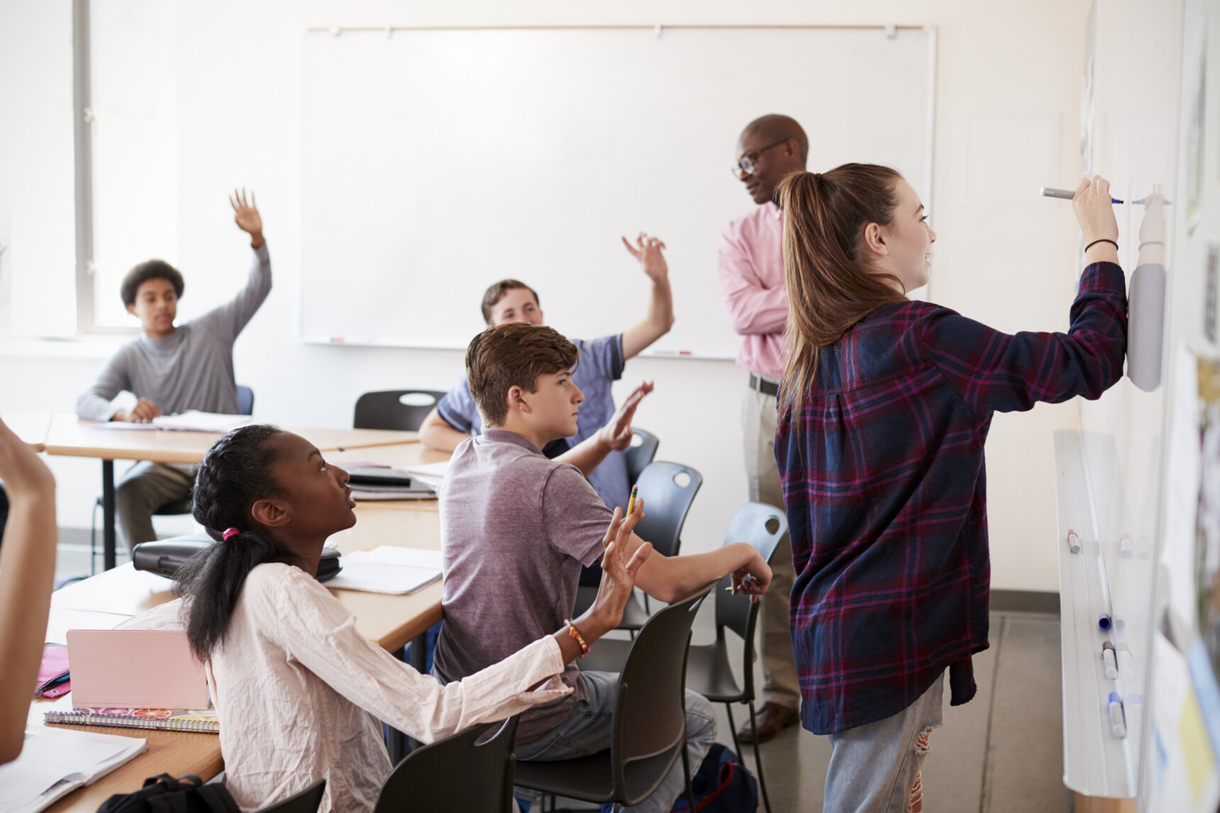 A classroom of students, where one student is dual coding an explanation by drawing on the whiteboard and the other students raise their hands to answer the teacher's question.
